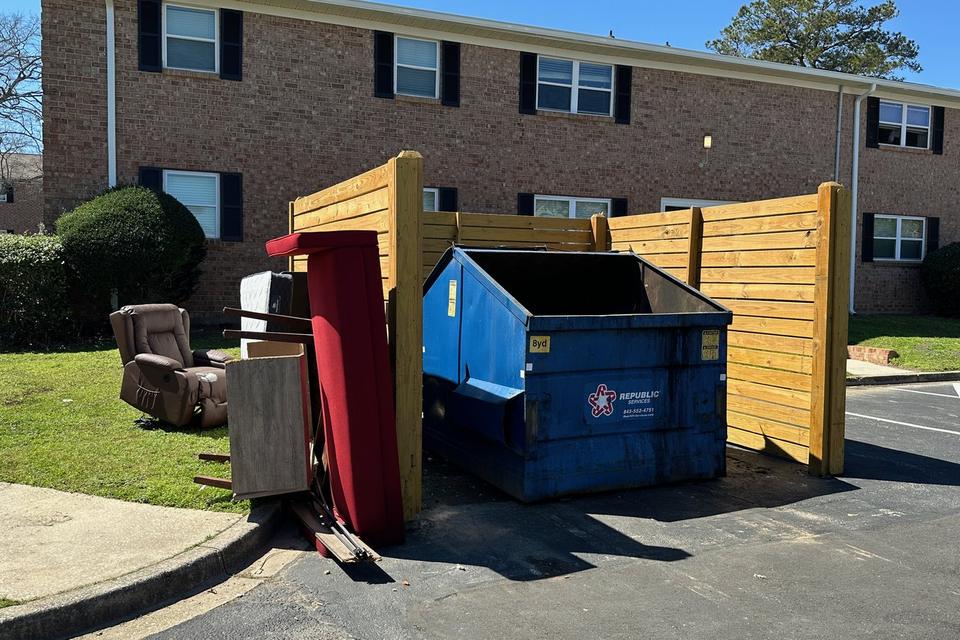 Dumpster corral at an apartment community with a recliner, mattress, and furniture piled beside it